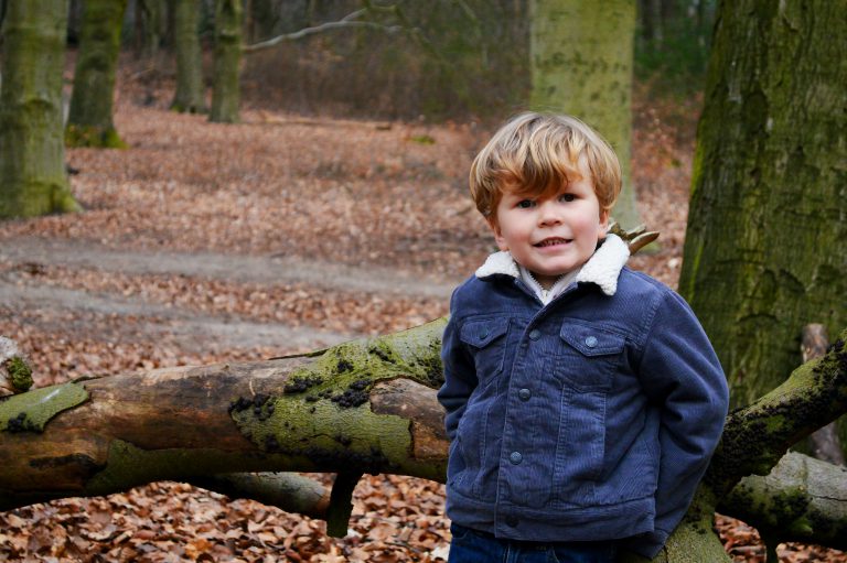 Small boy leaning against a tree with the woods behind him.